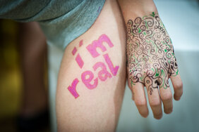 Das Bild zeigt eine Nahaufnahme eines Oberschenkels mit den Worten „I’m real“ in pinker Schrift und einer Hand mit kunstvoller, filigraner Bemalung in schwarzen und grünen Mustern, die an Henna-Tattoos erinnern.