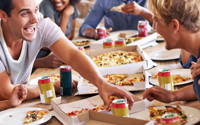 ine fröhliche Gruppe junger Erwachsener sitzt lachend an einem Holztisch und teilt sich mehrere Pizzen aus Pappkartons sowie Getränkedosen.