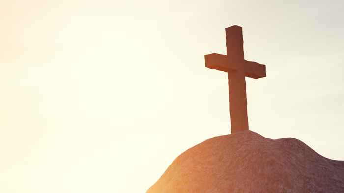 Ein Kreuz steht auf einem Hügel, beleuchtet von warmem Sonnenlicht am Horizont. @Canva