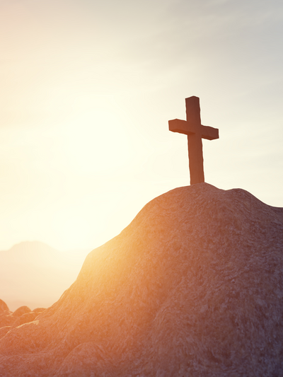 Ein Kreuz steht auf einem Hügel, beleuchtet von warmem Sonnenlicht am Horizont.