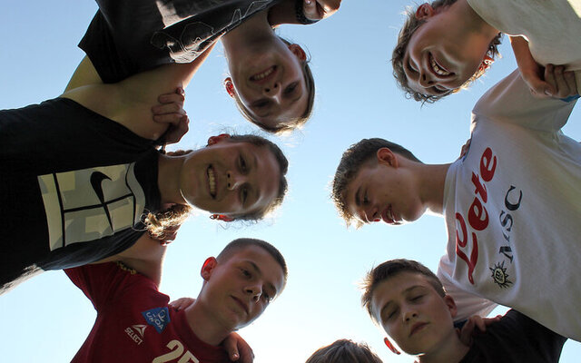 Das Bild zeigt eine Gruppe von Jugendlichen, die in einem Kreis stehen und sich die Arme um die Schultern legen. Sie schauen gemeinsam in die Kamera, die aus der Mitte des Kreises nach oben gerichtet ist. Der Himmel ist klar und blau im Hintergrund, was eine fröhliche und verbundene Atmosphäre vermittelt.