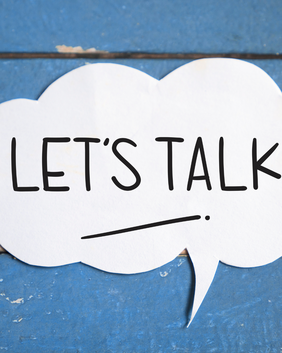 Ein weißes Sprechblasen-Ausschnitt mit der Aufschrift „LET’S TALK“ (Lass uns reden) liegt auf einem blauen, strukturierten Holzhintergrund.
