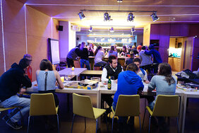 Das Bild zeigt eine Gruppe junger Menschen, die in einer gemütlichen und modernen Umgebung zusammenkommen. Im Vordergrund sitzen mehrere Personen an Tischen und spielen Brettspiele oder unterhalten sich. Im Hintergrund versammeln sich weitere Menschen an einer Bar oder Theke, was auf eine gemeinschaftliche Veranstaltung oder ein Treffen hindeutet. Die Beleuchtung mit LED-Streifen und Spots verleiht dem Raum eine warme, freundliche Atmosphäre.