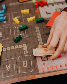 Ich überlege Eine Nahaufnahme zeigt die Hände einer Person, die orangefarbene Spielkarten von einem Spielbrett nimmt, auf dem sich bunte Spielfiguren befinden.