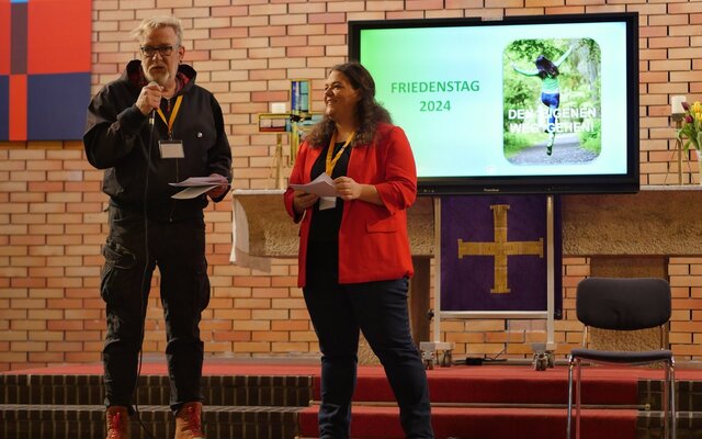 Das Bild zeigt zwei Personen auf einer Bühne in einem Kirchenraum. Eine Person, links im Bild, hält ein Mikrofon und spricht, während die andere Person, rechts, ein Blatt Papier in den Händen hält und lächelt. Im Hintergrund ist ein Bildschirm zu sehen, auf dem „Friedenstag 2024“ und das Bild einer Person in Bewegung mit dem Schriftzug „Den eigenen Weg gehen“ angezeigt werden. Die Szene wirkt wie eine Präsentation oder ein Vortrag im Rahmen einer Veranstaltung.